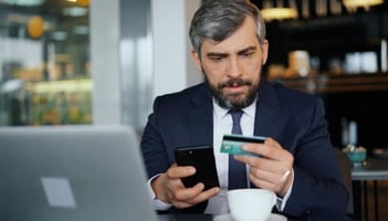 concerned professional middle-aged man looking at his credit card, holding a phone.