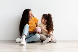 Smiling, young Hispanic woman and child holding a pink piggy bank, sitting on a floor, with their backs against a wall
