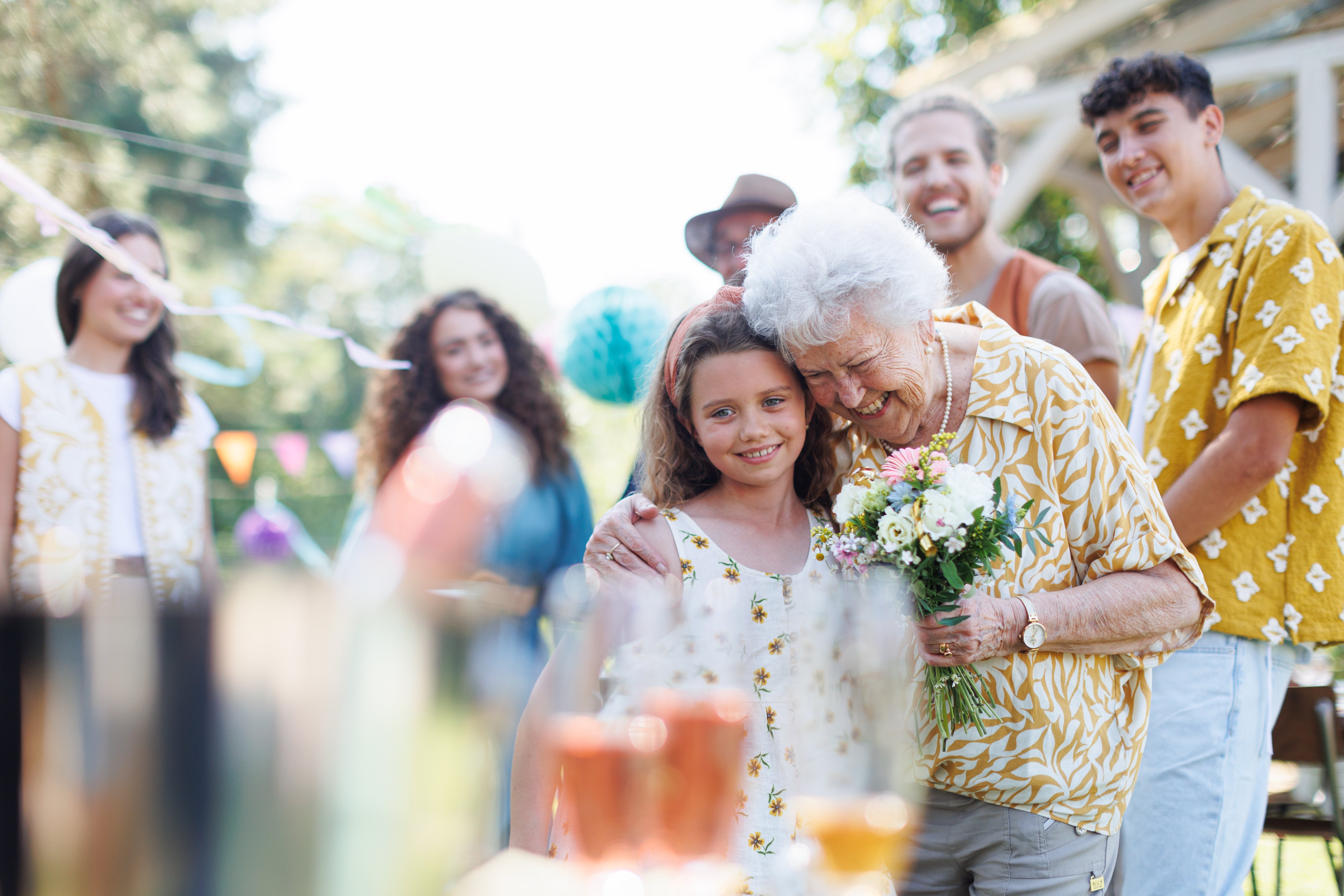grandmother hugging a young girl who just gave her a bouquet of flowers while several young adults watch in the background.