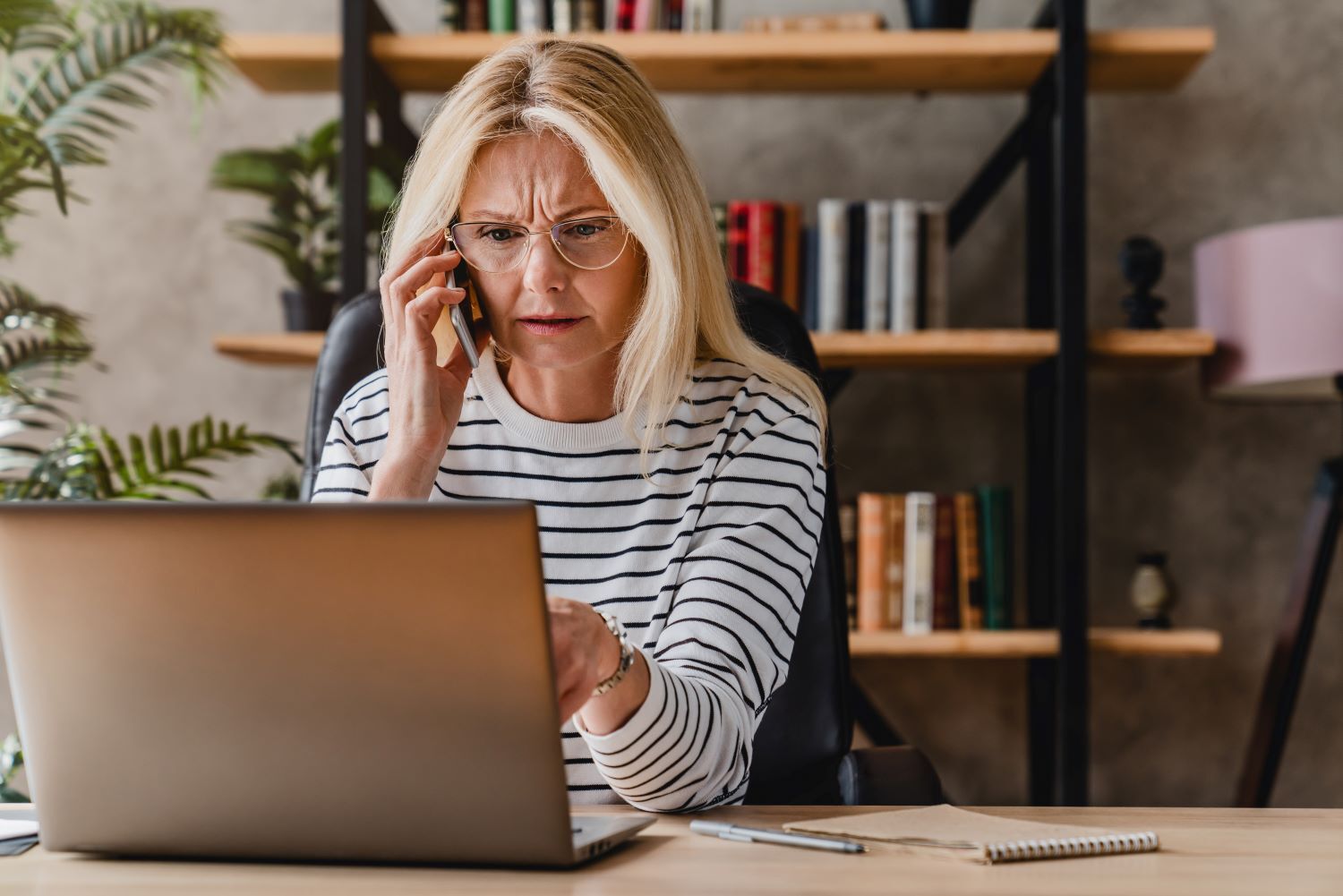 Distressed looking middle-aged blonde woman talking on the phone and looking at a laptop screen