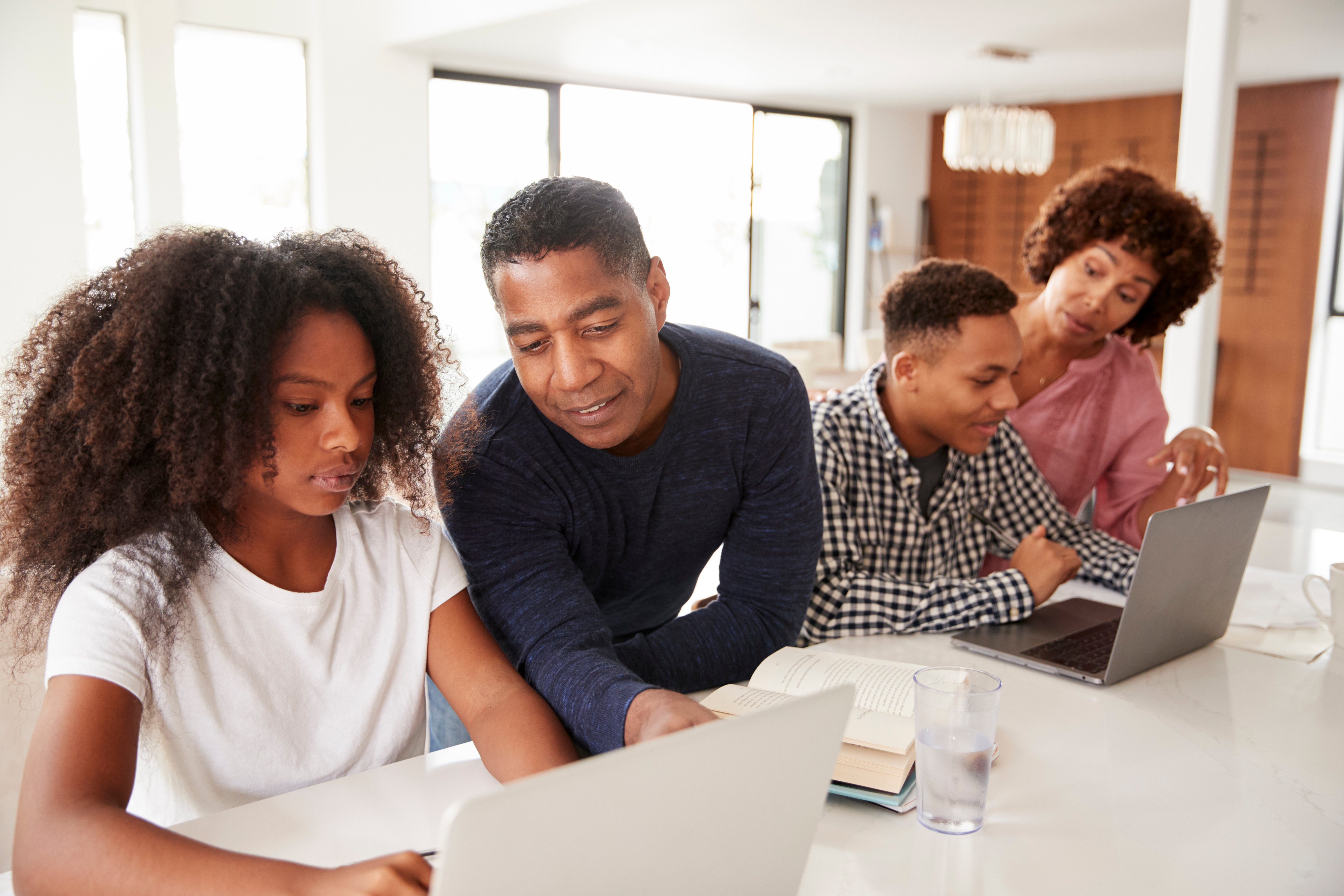middle aged black couple in a kitchen with their teen-aged daughter and son looking at computer screens.