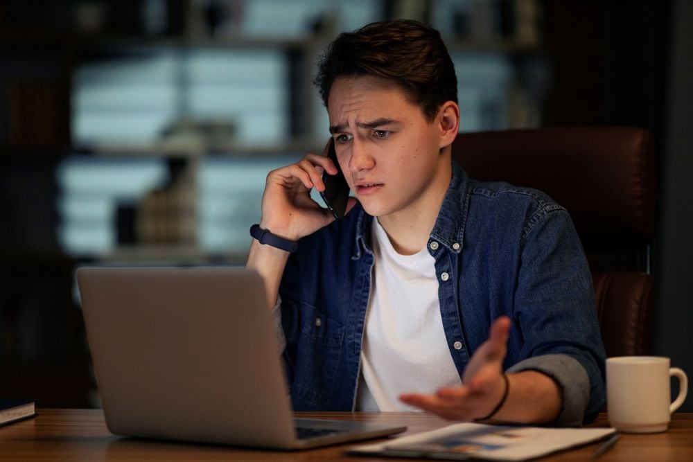 Young man looking at his computer and talking to his banker about being scammed. 